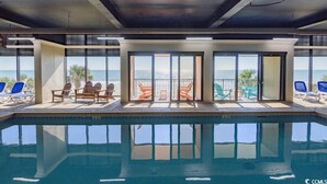 Indendørs swimmingpool med frit udsyn til havet gennem store vinduer, omgivet af liggestole.