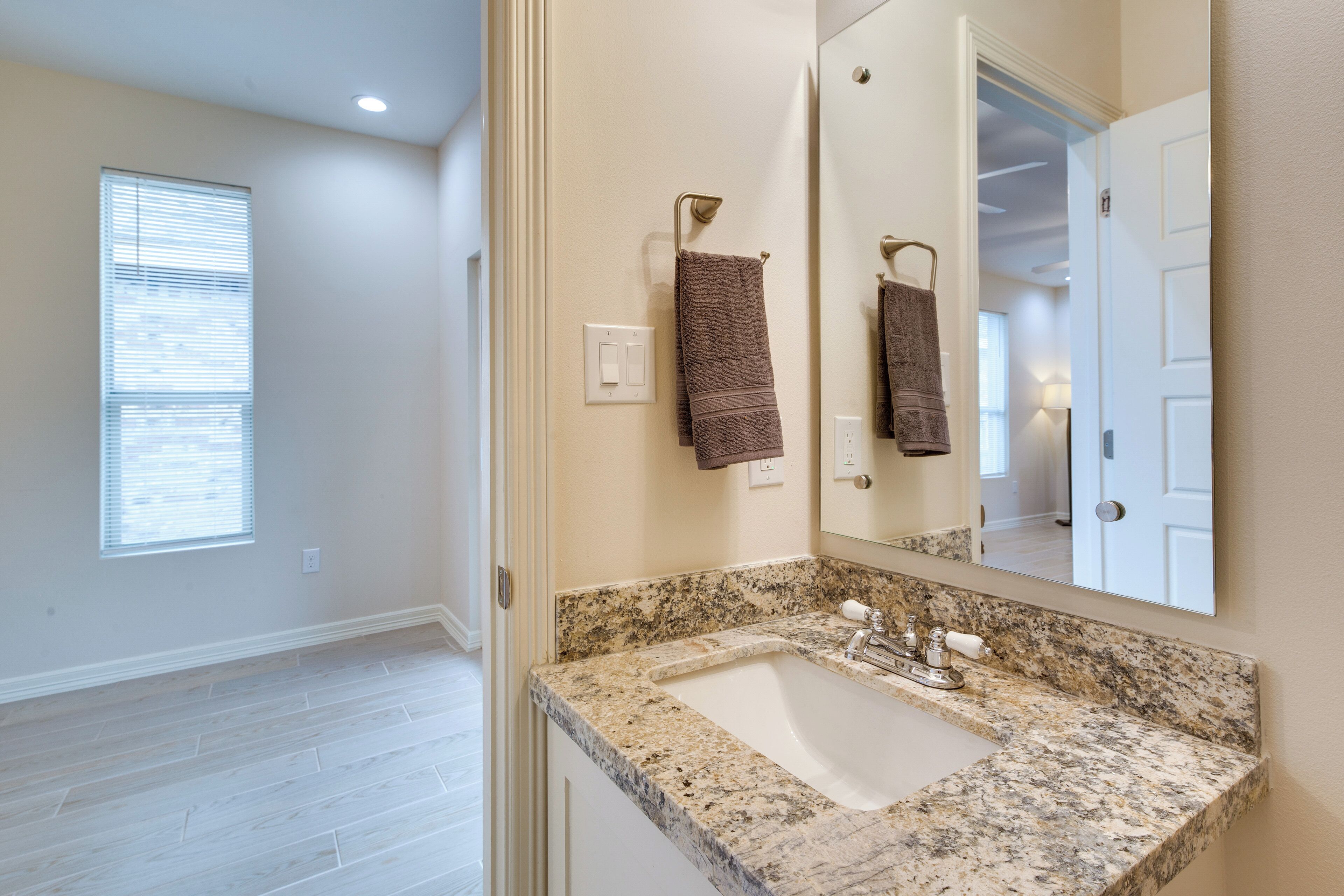 A bathroom with a granite countertop sink, a towel rack with two towels, a mirror, and a window with blinds.