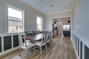 A dining area with a long wooden table and six chairs, a kitchen in the background, and a view of a brick building outside. - Modern Natchitoches Retreat on Cane River! (Natchitoches)
