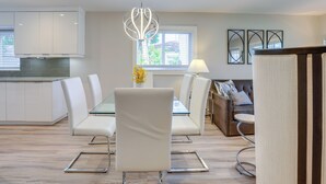 Uma cozinha moderna com armários brancos, um espaço de refeições com mesa de vidro e cadeiras brancas, e uma sala de estar com sofá e luminária.