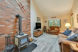Un salon chaleureux avec un mur en briques, un poêle à bois, un téléviseur posé sur un meuble en bois, un canapé en cuir marron et une fenêtre donnant sur les arbres.
