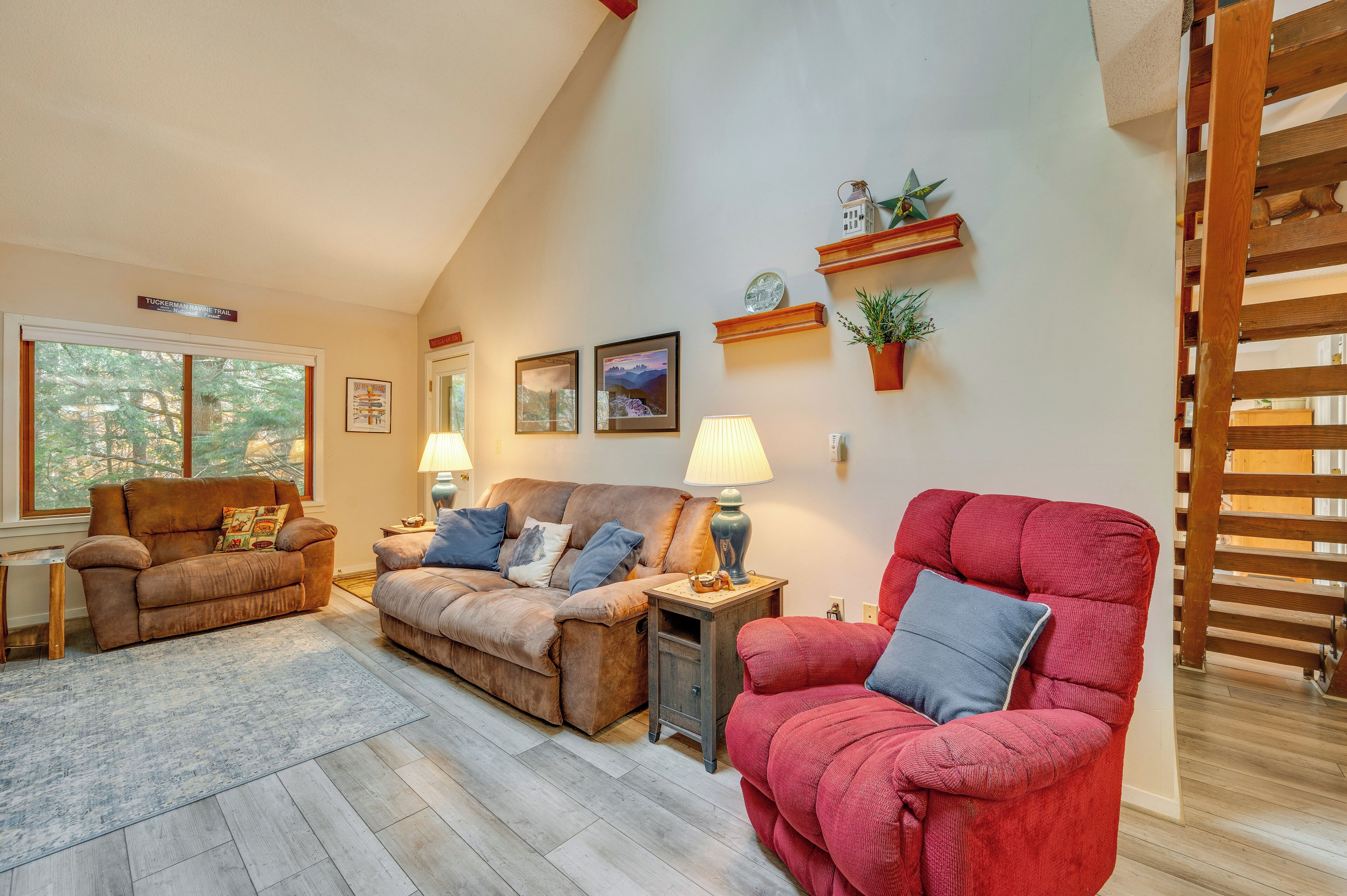 A cozy living room with a brown sofa, a red armchair, and a wooden coffee table. There are two wall-mounted lamps and a small shelf with decorative items.