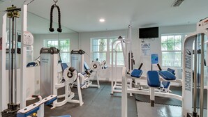 Un gimnasio con diversas máquinas de ejercicio, un televisor montado en la pared y amplios ventanales que dejan entrar luz natural.