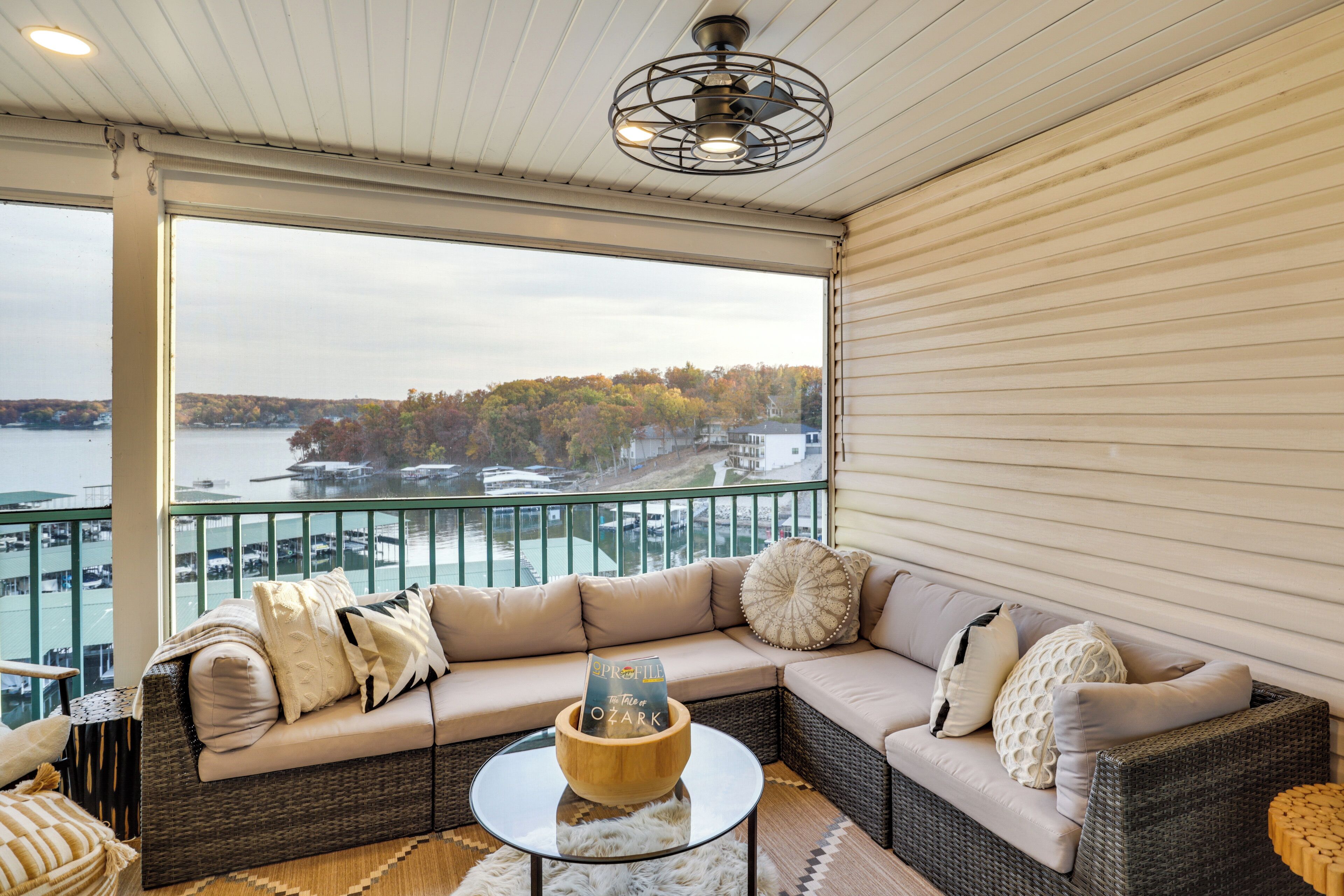 Un balcon avec un canapé en osier, une table basse en verre et une vue sur un lac et des arbres.