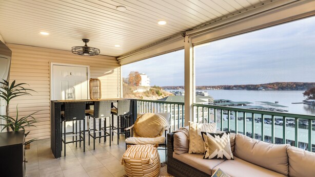 A balcony with a view of a body of water and a distant bridge. There is a seating area with a sofa and chairs, and a small bar area with stools and a refrigerator.