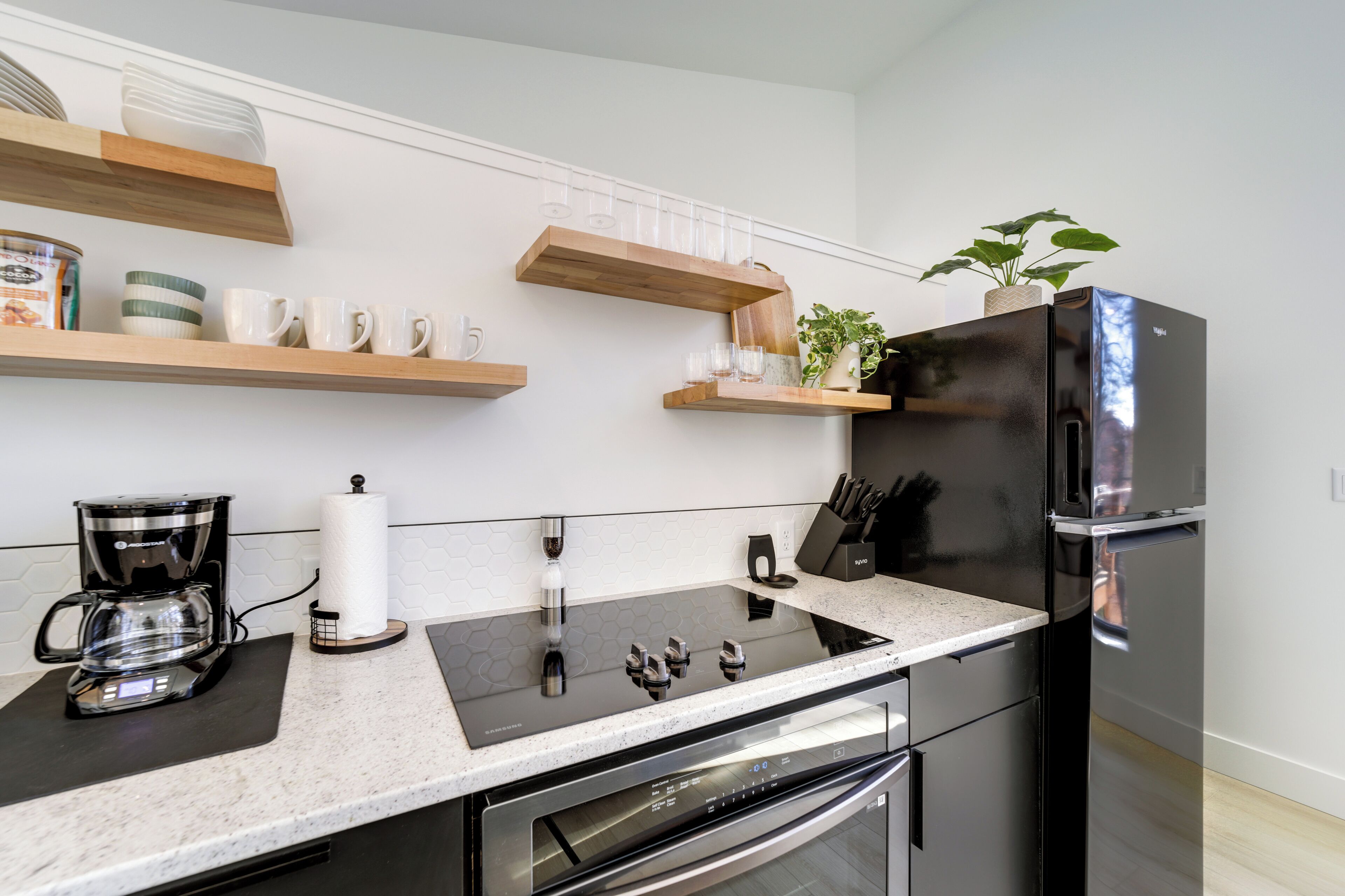 A modern kitchen with a built-in stove, oven, and refrigerator, wooden shelves, and a countertop with a coffee maker and glassware.