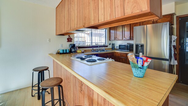 A kitchen with wooden cabinets, a stainless steel refrigerator, a microwave, and a granite countertop. There are two stools and a basket of snacks on the counter. - Adobe Home in Albuquerque w/ Covered Patio (Albuquerque)