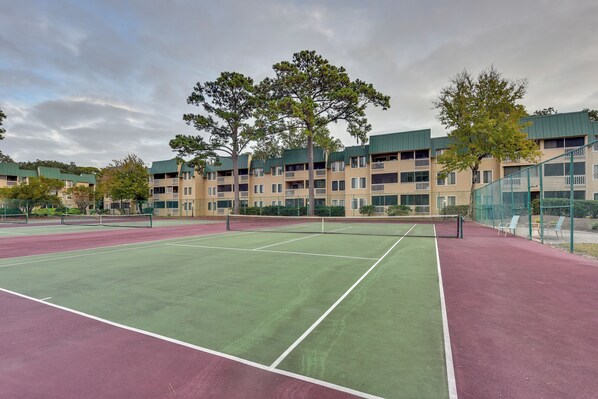 A tennis court with a net, surrounded by multi-story buildings with balconies and trees.