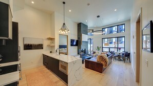 A modern kitchen with a marble island, a dining area with a sofa and table, and a large window providing natural light. - Luxe Schweitzer Condo w/ Hot Tub: Ski-in/ski-out! (Sandpoint)
