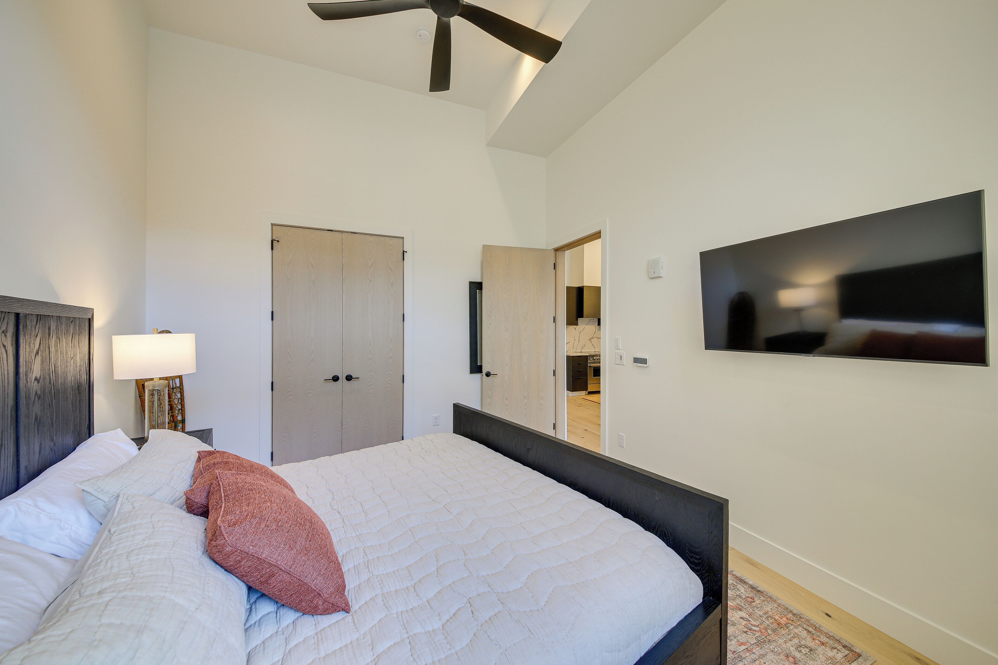 A bedroom with a bed, a ceiling fan, a TV mounted on the wall, and a door leading to another room.