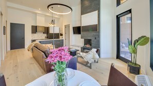 A modern living room with a sofa, a dining table with a vase of flowers, a fireplace, and a large window. - Luxe Schweitzer Condo w/ Hot Tub: Ski-in/ski-out! (Sandpoint)