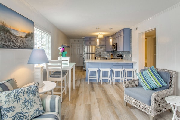 A modern kitchen with a central island, bar stools, and a wicker sofa with a striped pillow. - Steps to Ocean: High-end North Myrtle Beach Condo (North Myrtle Beach)