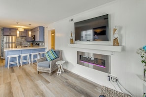 A modern kitchen with a bar area, a flat-screen TV mounted on the wall, and a fireplace below it. - Steps to Ocean: High-end North Myrtle Beach Condo (North Myrtle Beach)