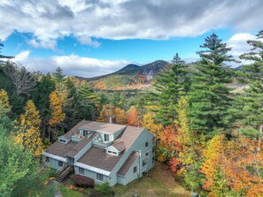 Une maison avec un toit en pente, entourée d’un feuillage automnal et avec des montagnes en arrière-plan.