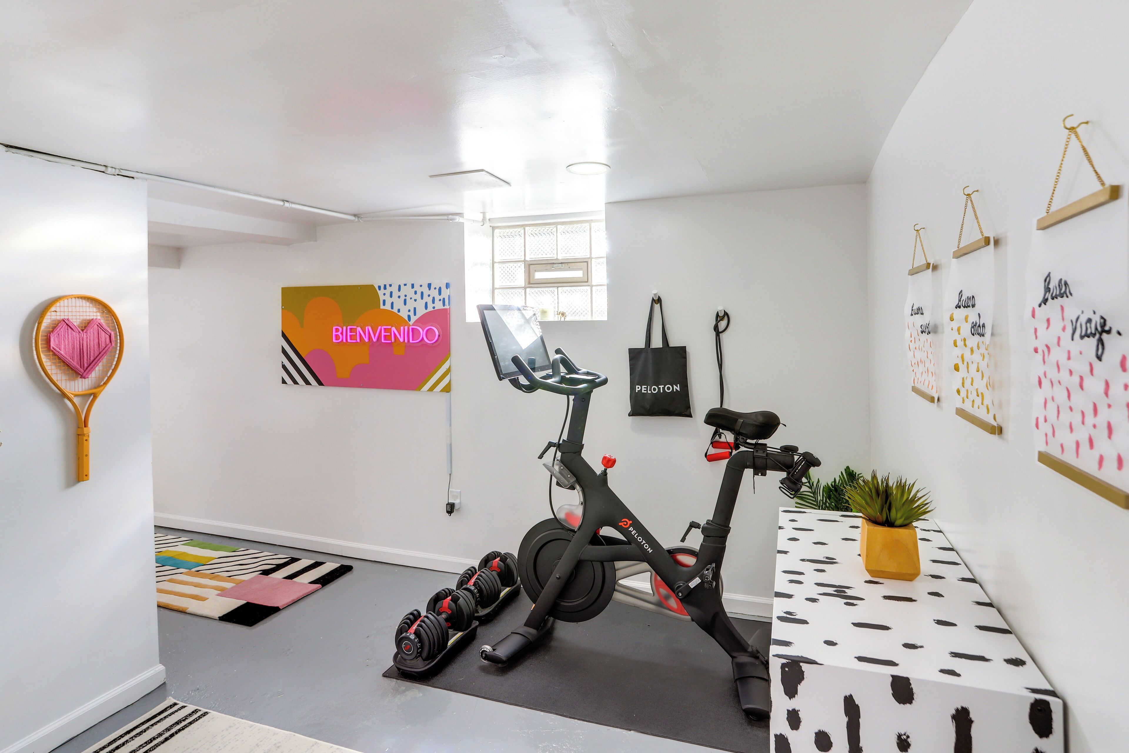 Une pièce moderne et bien éclairée, équipée d’un vélo d’appartement, d’un tapis coloré et d’un sac Peloton fixé au mur.