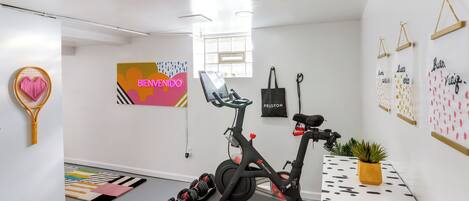 Une pièce moderne et bien éclairée, équipée d’un vélo d’appartement, d’un tapis coloré et d’un sac Peloton fixé au mur.