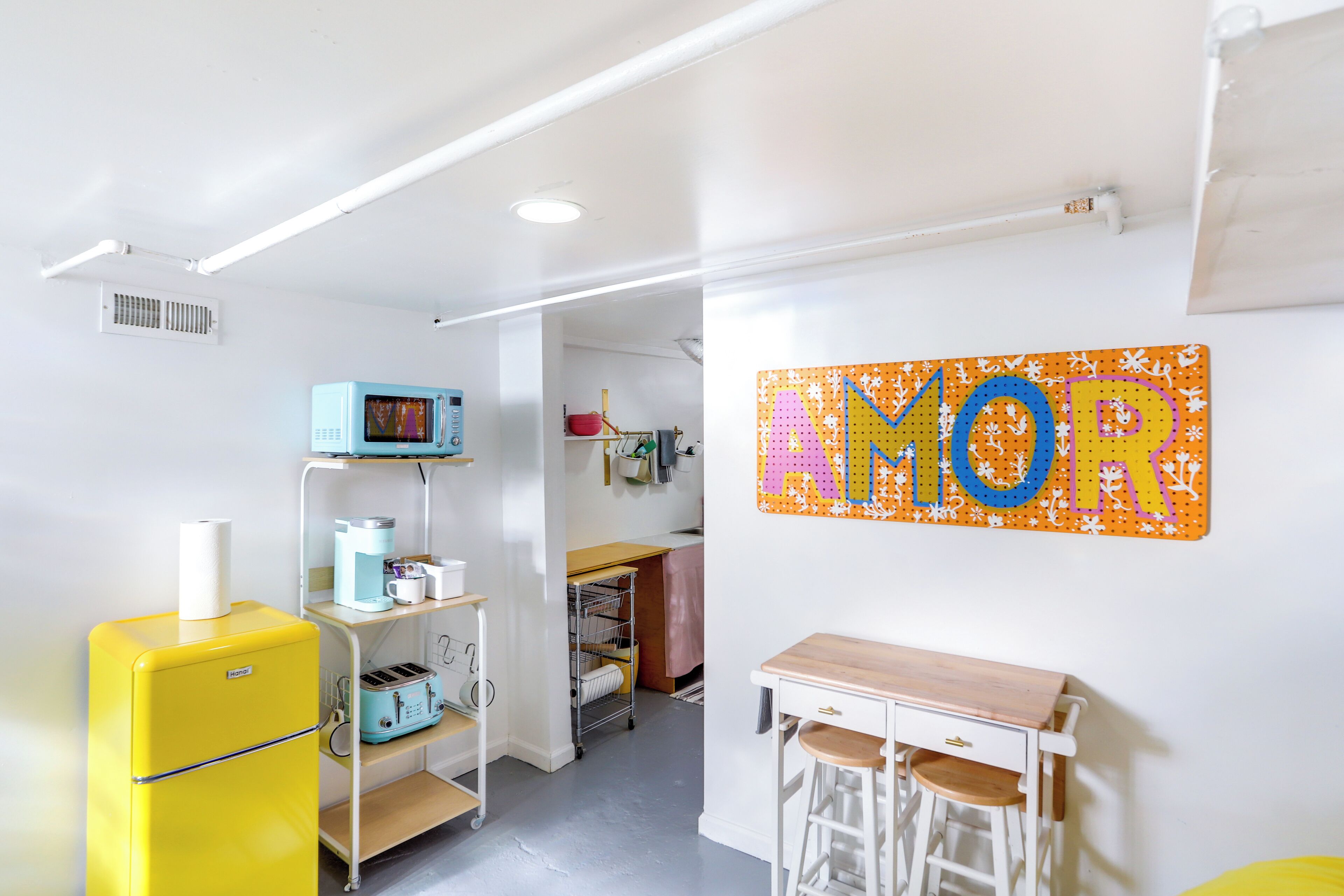 Una habitación con refrigerador amarillo, microondas, una mesita con taburetes y un letrero en la pared que dice 'AMOR'.