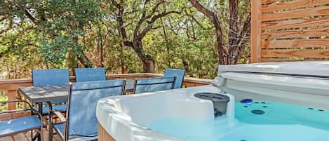 A hot tub on a wooden deck with chairs and a table, surrounded by trees.