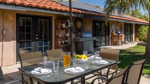 An outdoor dining area with a table set for four, a grill, and a sun dial on the wall. - La Buena Vida – Palm Springs Pool & Spa Retreat (Palm Springs)