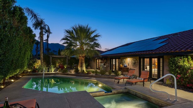 A swimming pool with lounge chairs and a table, surrounded by palm trees and a house with a tiled roof. - La Buena Vida – Palm Springs Pool & Spa Retreat (Palm Springs)