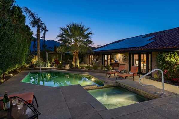 A swimming pool with lounge chairs and a table, surrounded by palm trees and a house with a tiled roof.