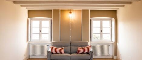 Una habitación amplia con un sofá gris, dos ventanas con marcos blancos y un radiador debajo de cada ventana.