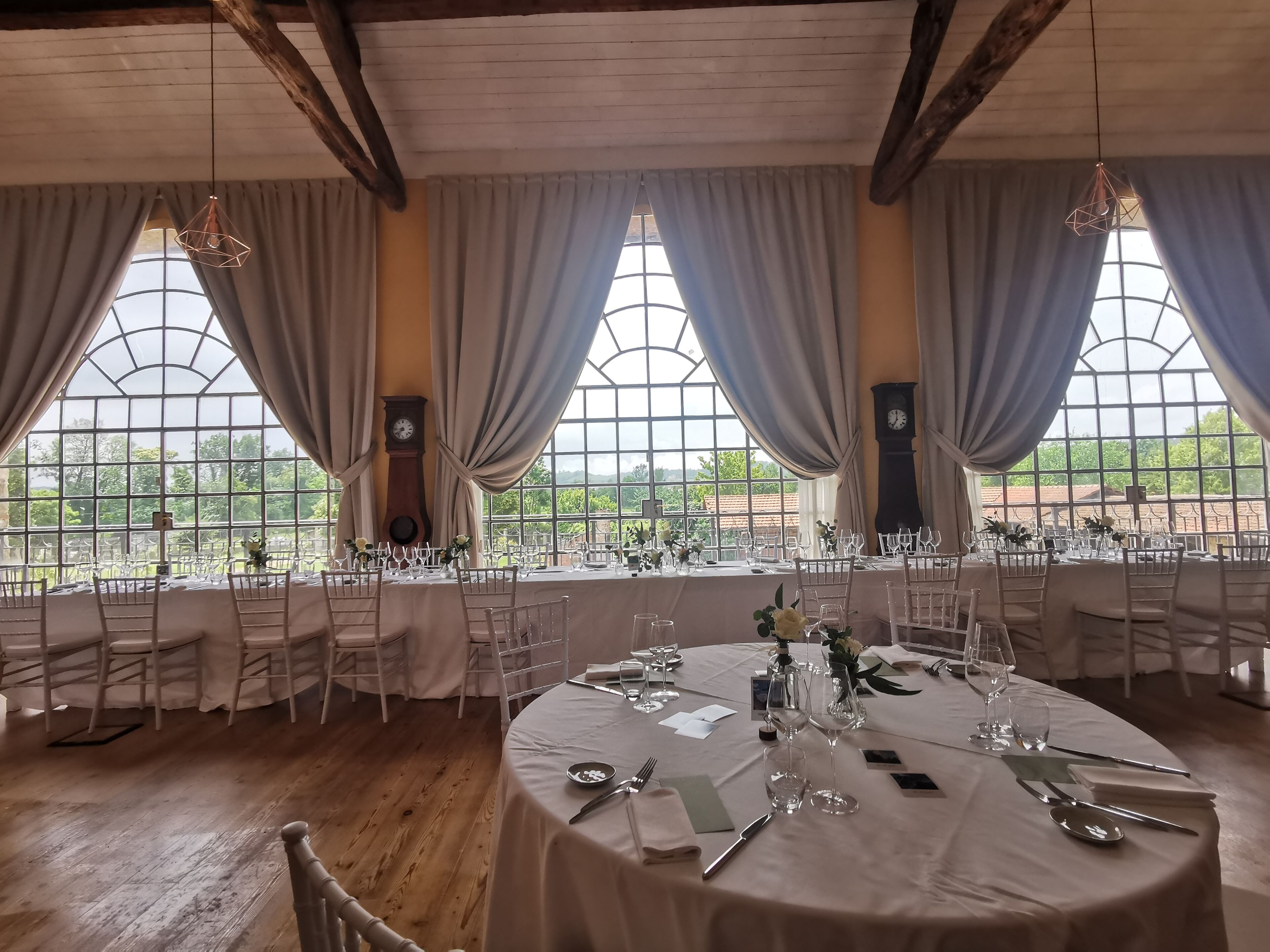 Un comedor con mesas redondas dispuestas para una comida, ventanas amplias con cortinas y vigas de madera en el techo.