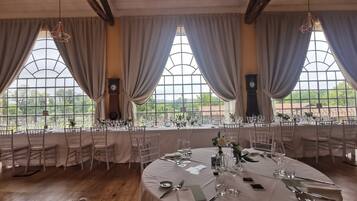 Un comedor con mesas redondas dispuestas para una comida, ventanas amplias con cortinas y vigas de madera en el techo.