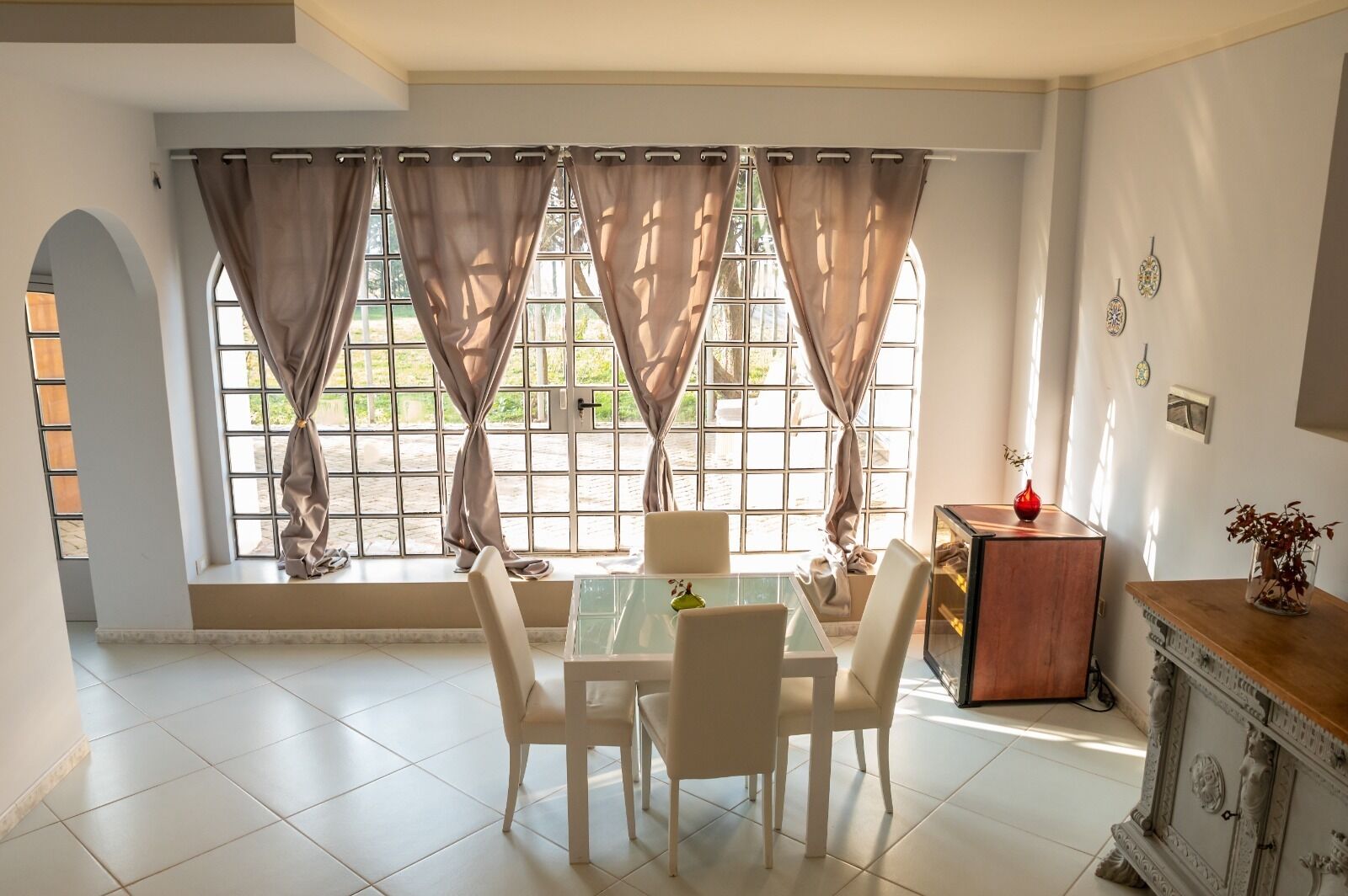 Un comedor con una mesa y sillas, una ventana con cortinas, un mueble aparador y un florero con flores.