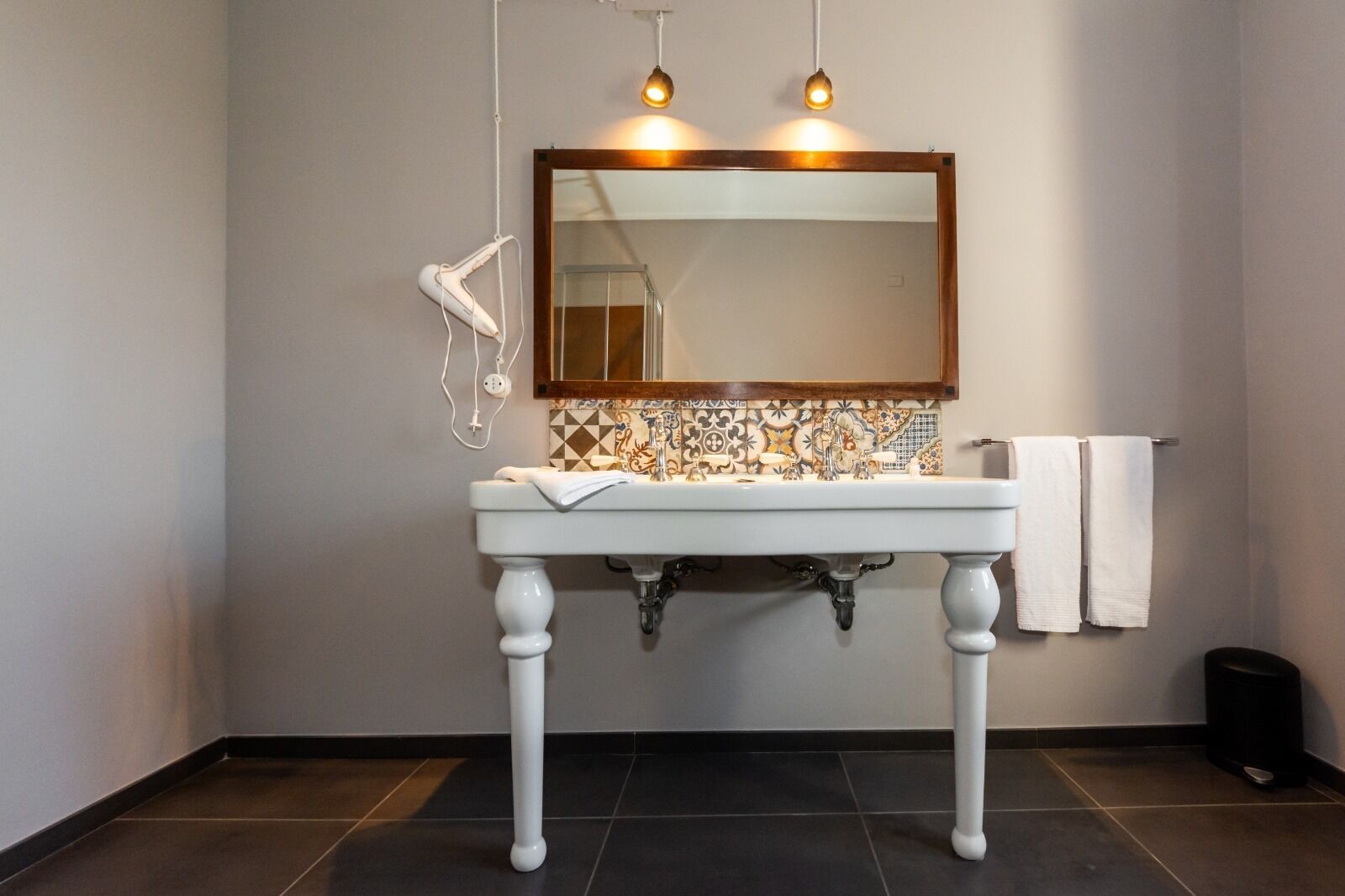 A bathroom with a white pedestal sink, a tiled backsplash, and a mirror above it. There are two white towels hanging on the wall and a hairdryer on the left.