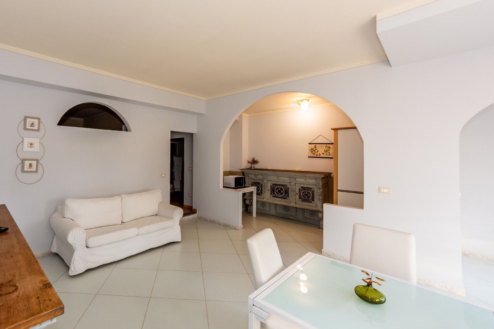 A modern living room with a white sofa, a glass dining table with chairs, and a kitchen area with a microwave and decorative cabinet.