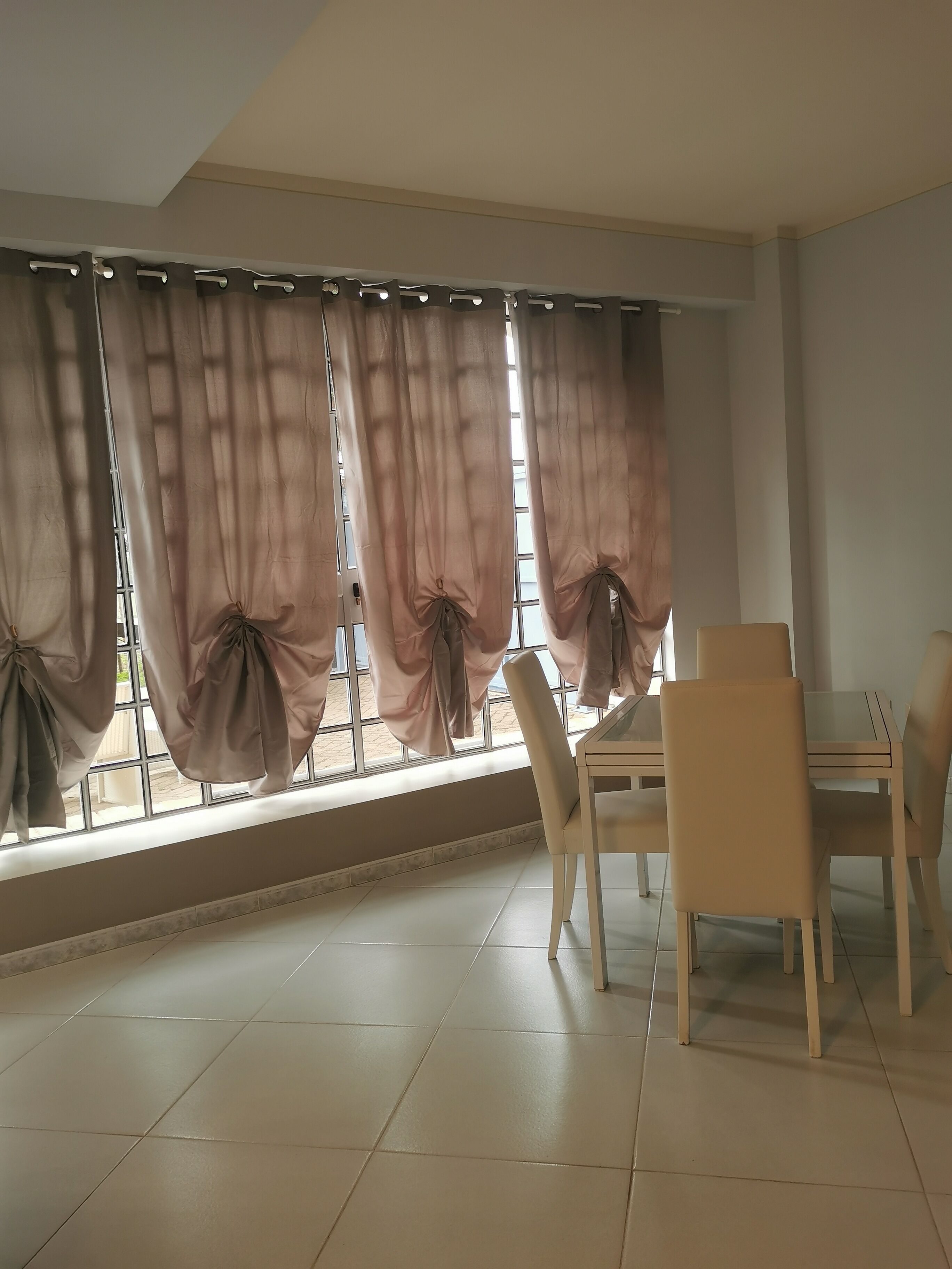 A dining area with a table and chairs, large windows with curtains, and a tiled floor.