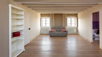 Una habitación amplia con un sofá gris, estanterías blancas y una ventana con radiador.