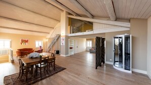A spacious loft with high ceilings, a dining area with a wooden table and chairs, a staircase leading to an upper level, and a modern glass-enclosed room.