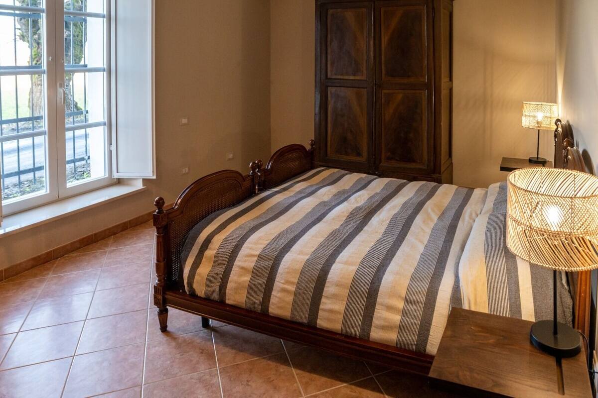 A bedroom with a wooden bed, striped bedding, a window, a cabinet, and a lamp.