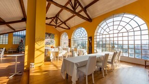 A dining area with high ceilings, wooden beams, and large arched windows. Tables are set with white tablecloths and chairs.