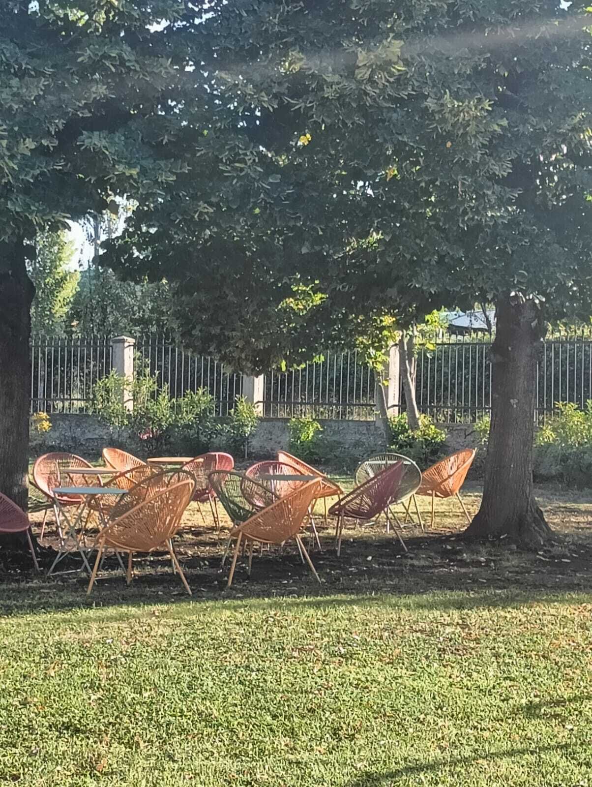 Buiten zitplaatsen met stoelen en tafels onder bomen, een metalen hek en een gazon.