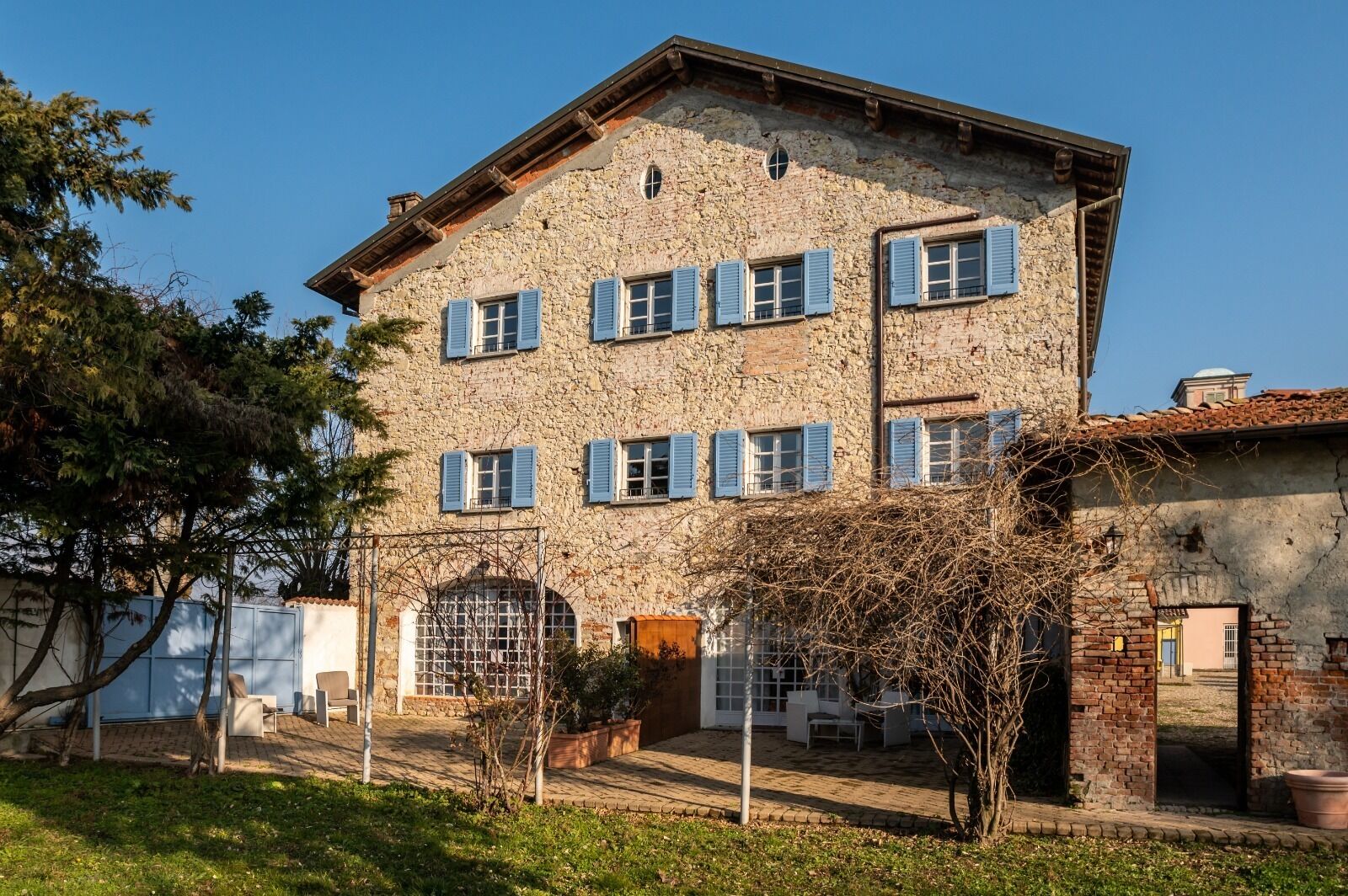Un bâtiment en pierre doté de nombreuses fenêtres et d’une porte, entouré d’un jardin et d’arbres.