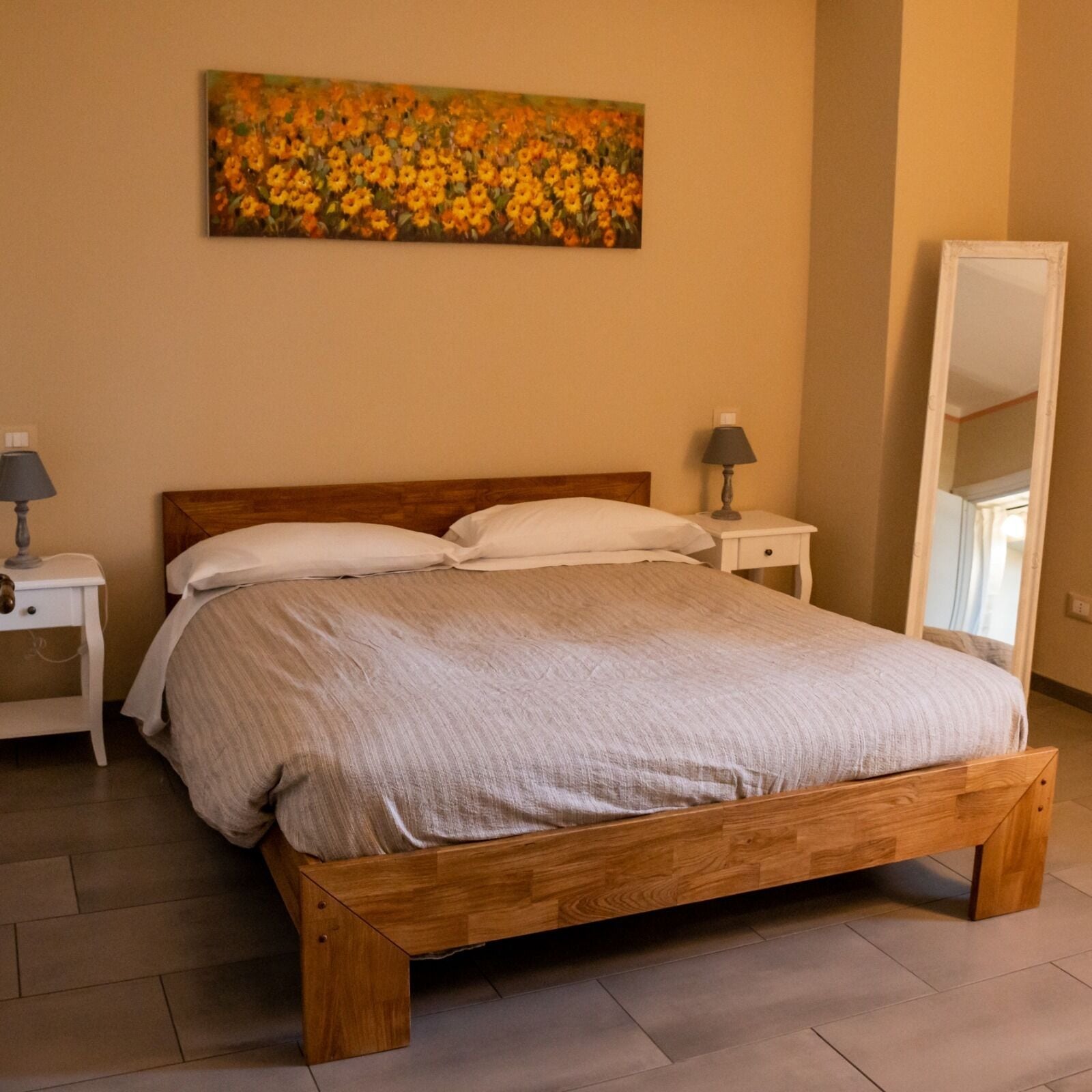 A bedroom with a wooden bed, white bedding, a nightstand with a lamp, a bedside table, and a large mirror.