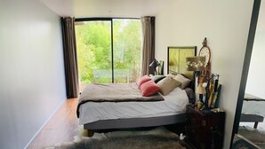 A bedroom with a large bed, a nightstand, a lamp, and a view of greenery outside.