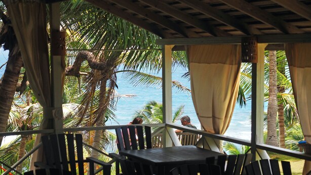 A covered outdoor seating area with tables and chairs, overlooking a beach with palm trees and the ocean. - Anmut (Dar es Salaam)