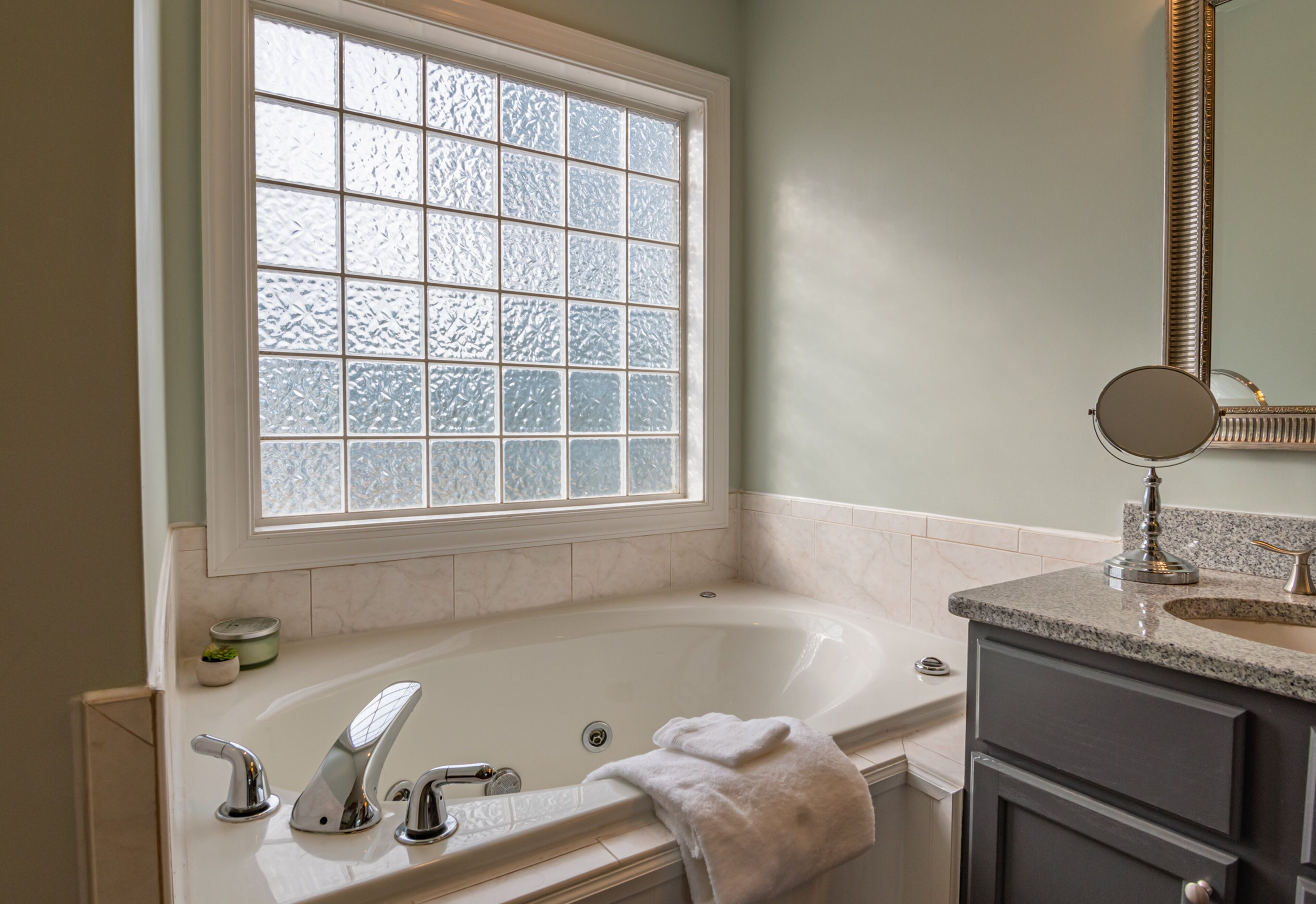 A bathroom with a large bathtub, a window with frosted glass, a sink with a granite countertop, and a mirror above the sink.