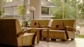 A modern living room with a sofa and chairs, a lamp, and a view of a house and trees through large windows. - Anmut (Dar es Salaam)