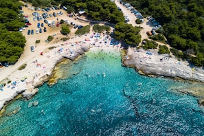 Luchtbeeld van een strand met helder turquoise water, rotsformaties en veel strandparasols en ligbedden.