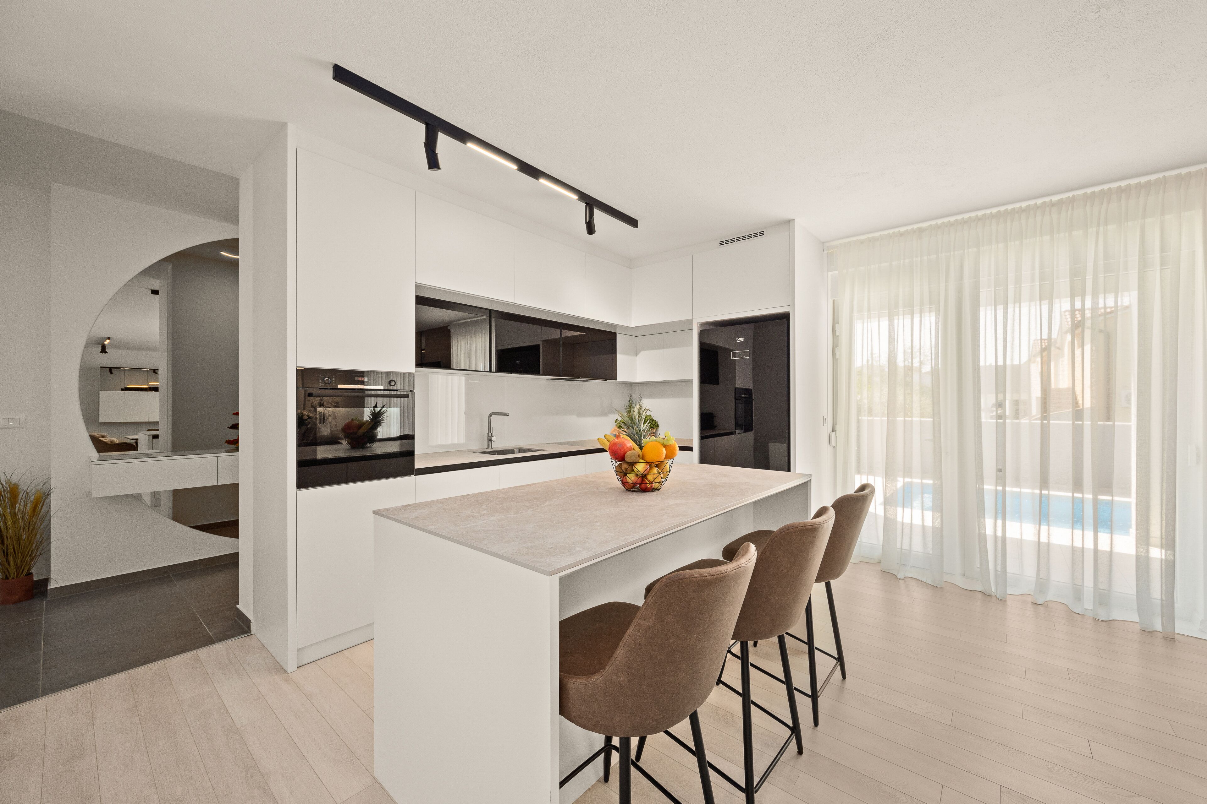A modern kitchen with a central island, bar stools, and a view of a pool through sheer curtains.