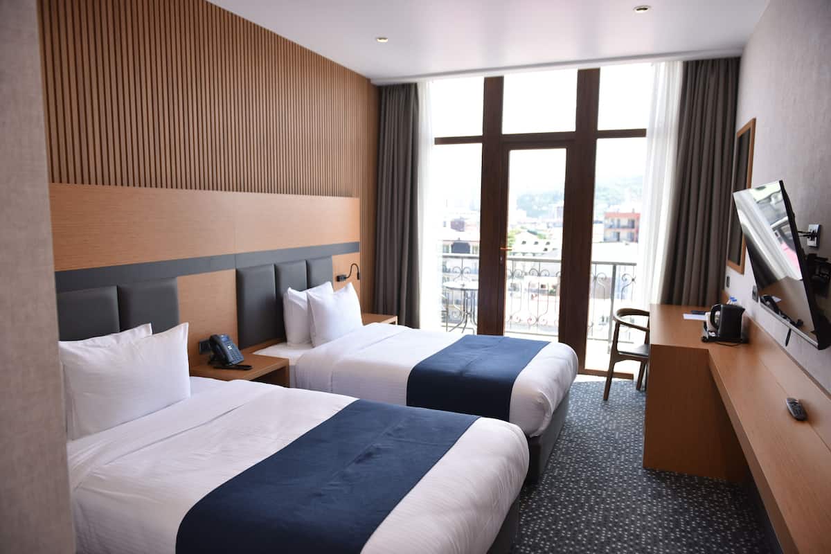 Habitación de hotel con dos camas, un escritorio, una silla, un televisor y un balcón con vista a la ciudad.