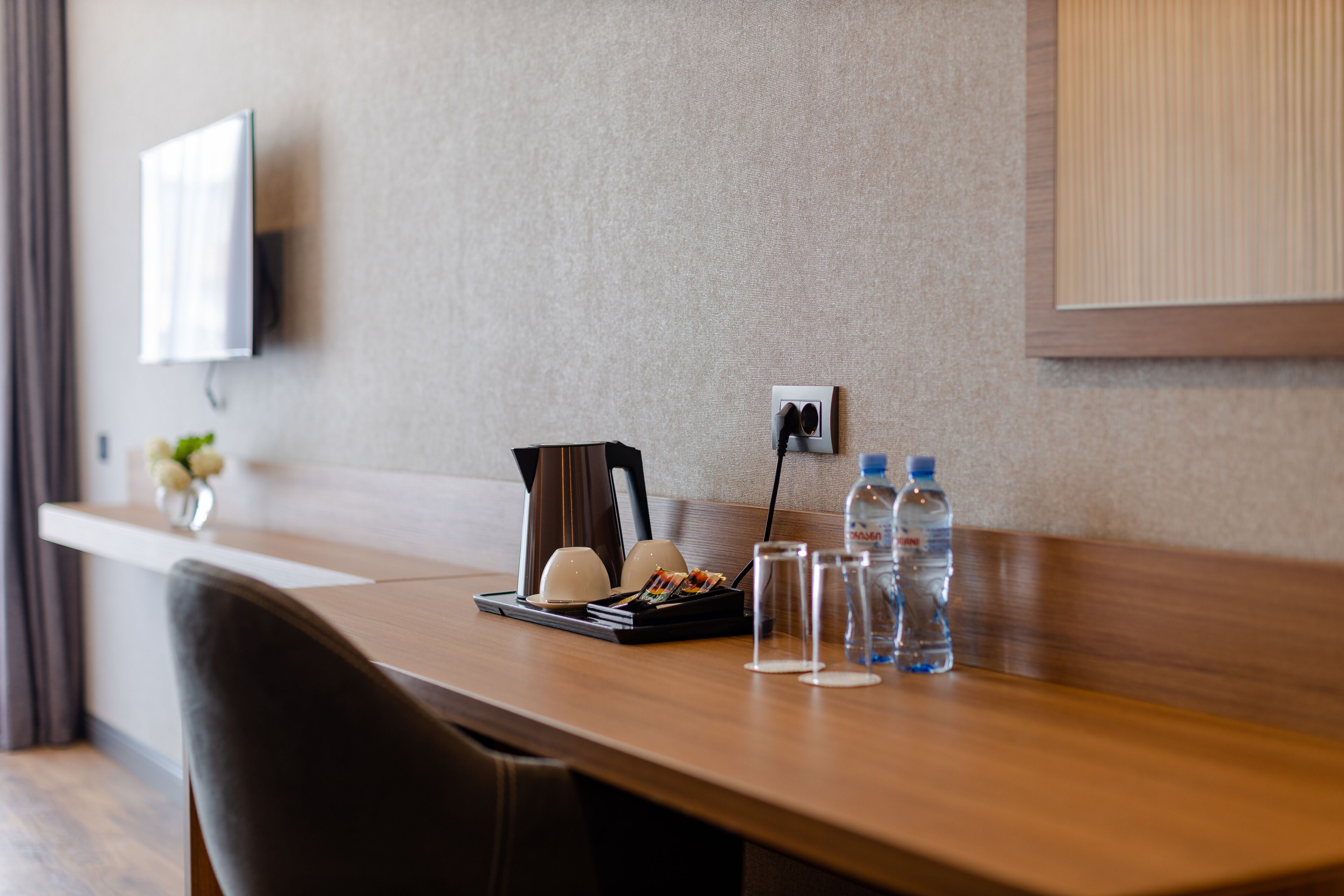 A hotel room with a wooden desk, a chair, a flat-screen TV mounted on the wall, a small table with a vase of flowers, and a water bottle.