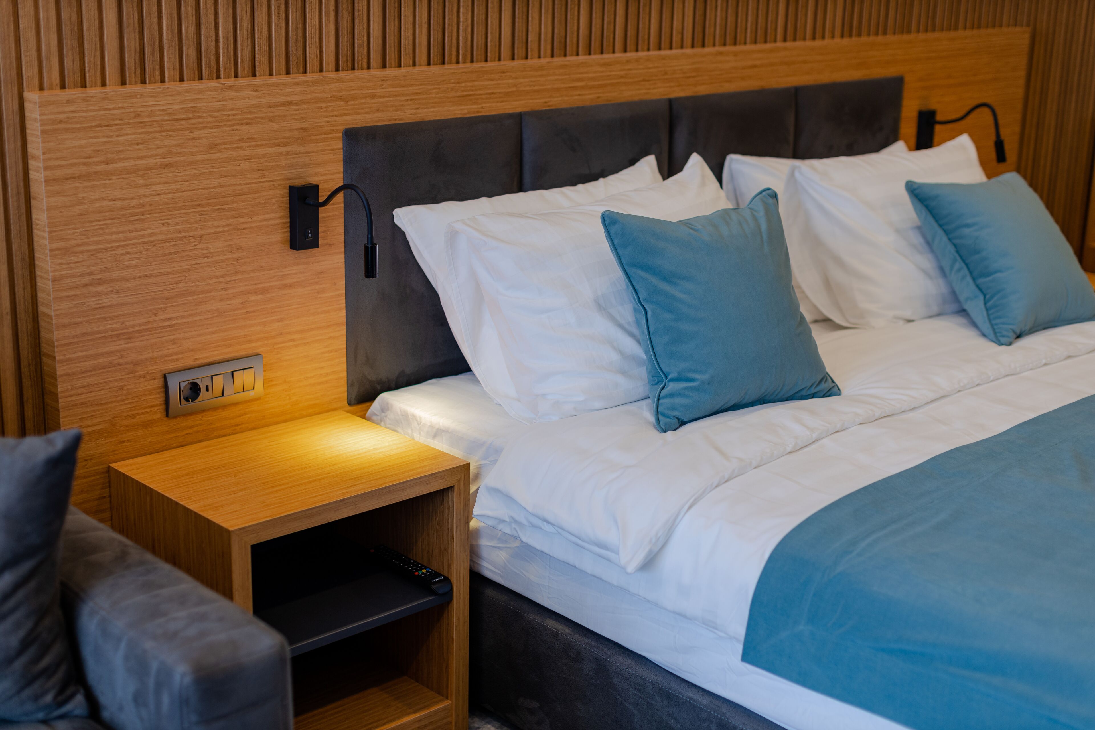 A neatly made bed with white and blue bedding, a wooden nightstand with a remote control drawer, and a wall-mounted light fixture.
