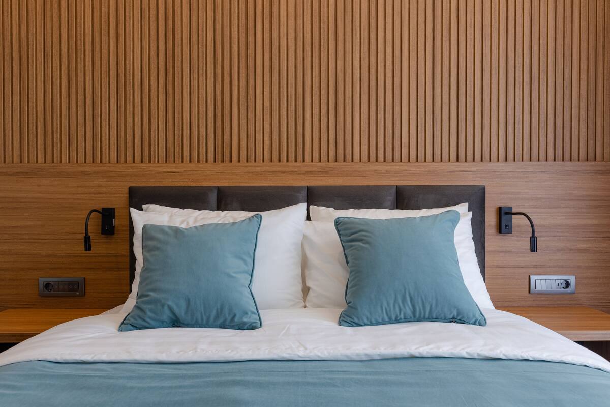 Una cama bien hecha con ropa de cama blanca y aguamarina, un cabecero de madera y dos mesitas de noche fijadas a la pared con lámparas.