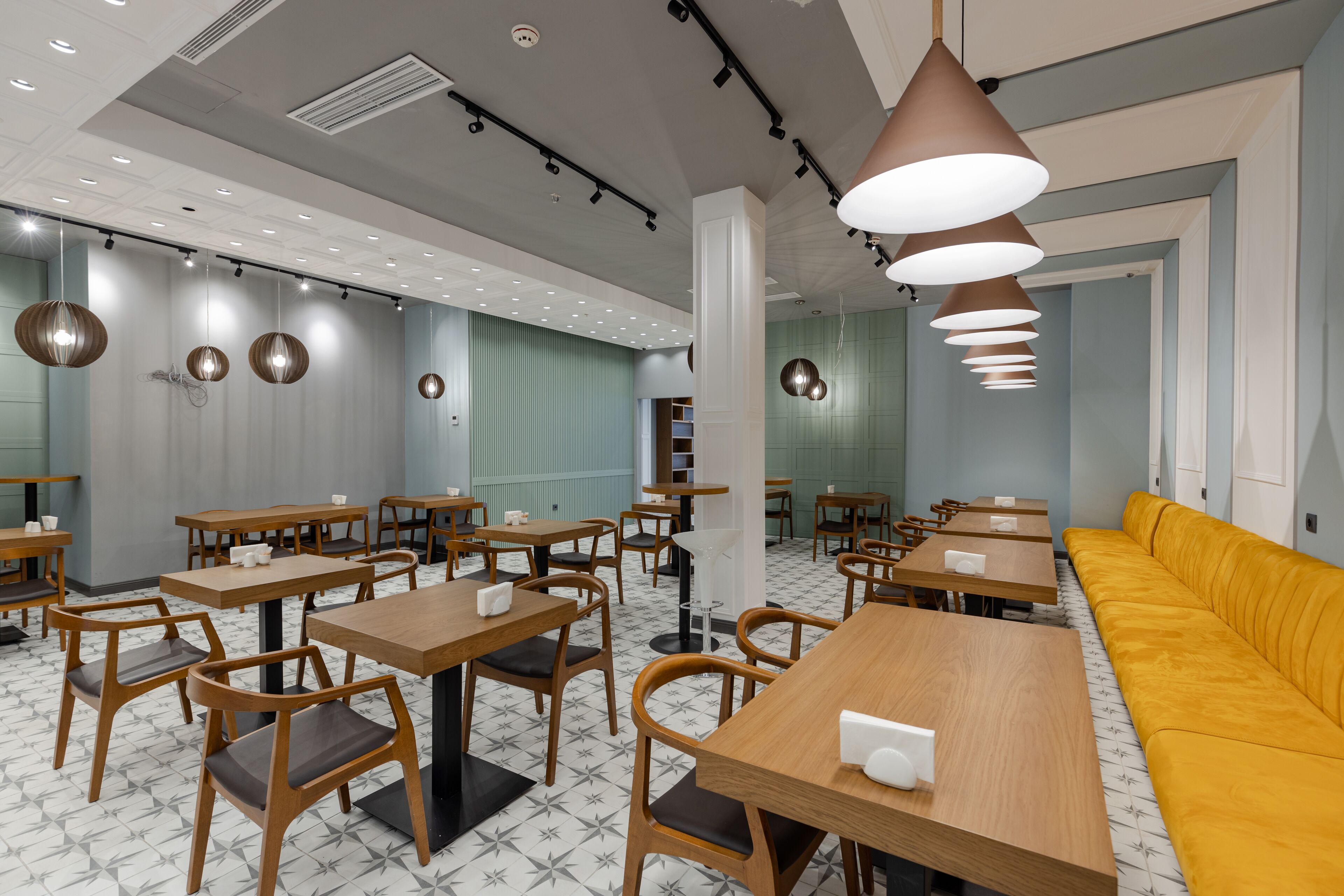 A modern cafe with wooden tables and chairs, a yellow sofa, and pendant lighting.